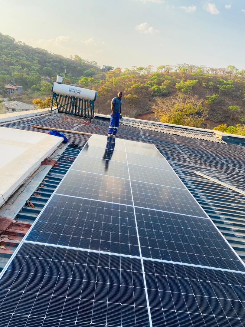 Dosmac two-man team commissioning large solar array on commercial rooftop, Harare hills in background