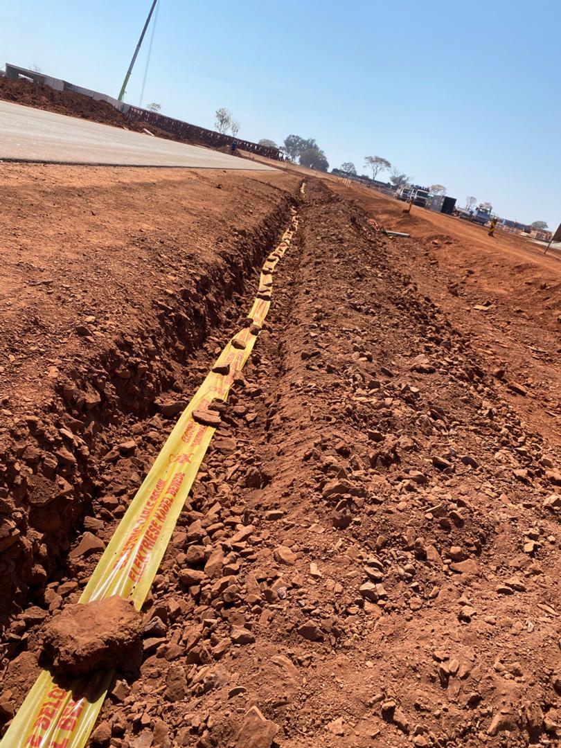 Underground armoured electrical cable laid in long straight trench with yellow warning tape, red Zimbabwe soil